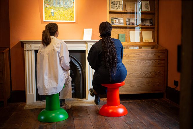 Picture of two people lisetning to soundscape of We Came Here. One woman is sat on a green stool, while the other woman is set on a red stool. Their backs are facing the camera and they are looking up.